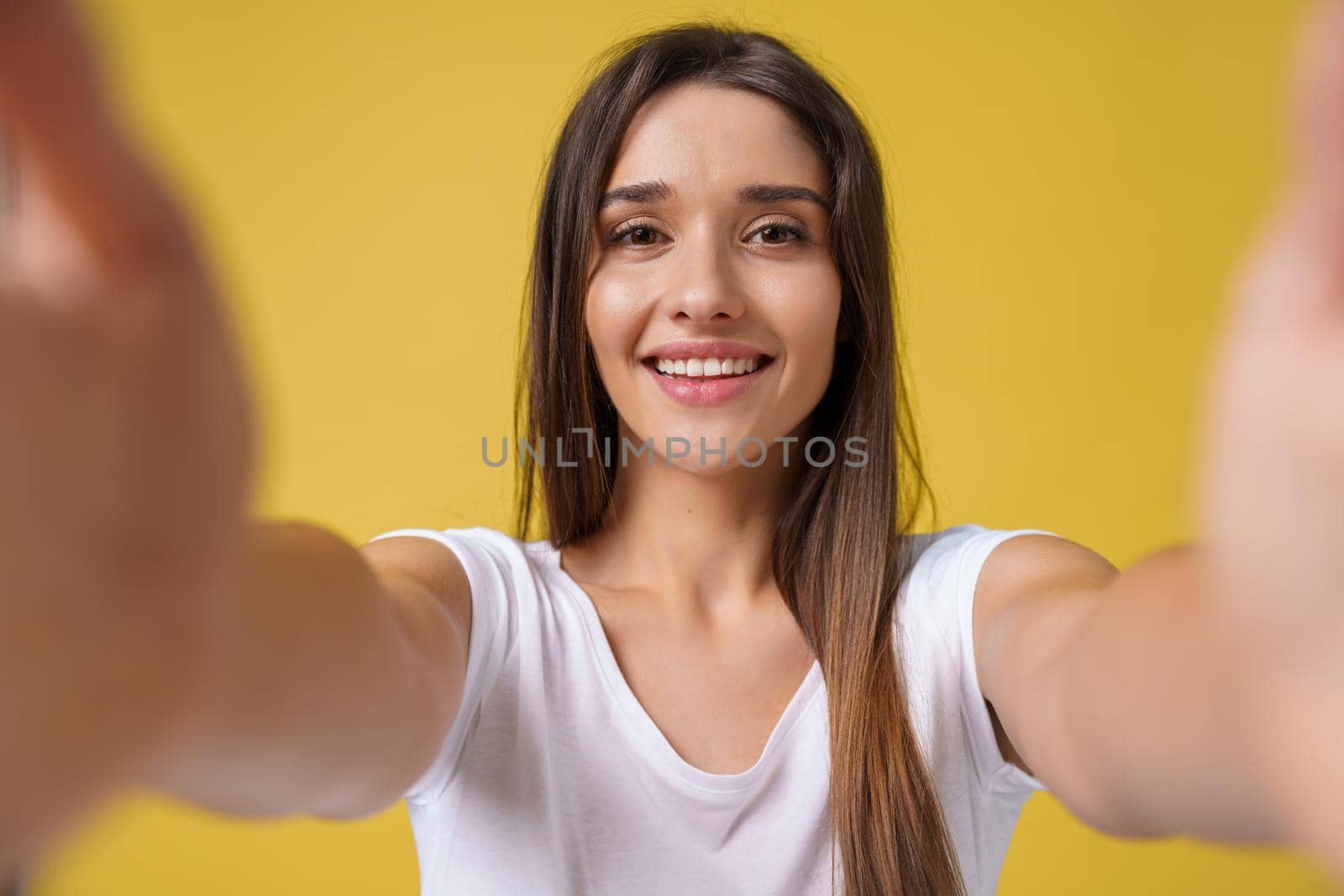 Pleasant attractive girl making selfie in studio and laughing. Good-looking young woman with brown hair taking picture of herself on bright yellow background