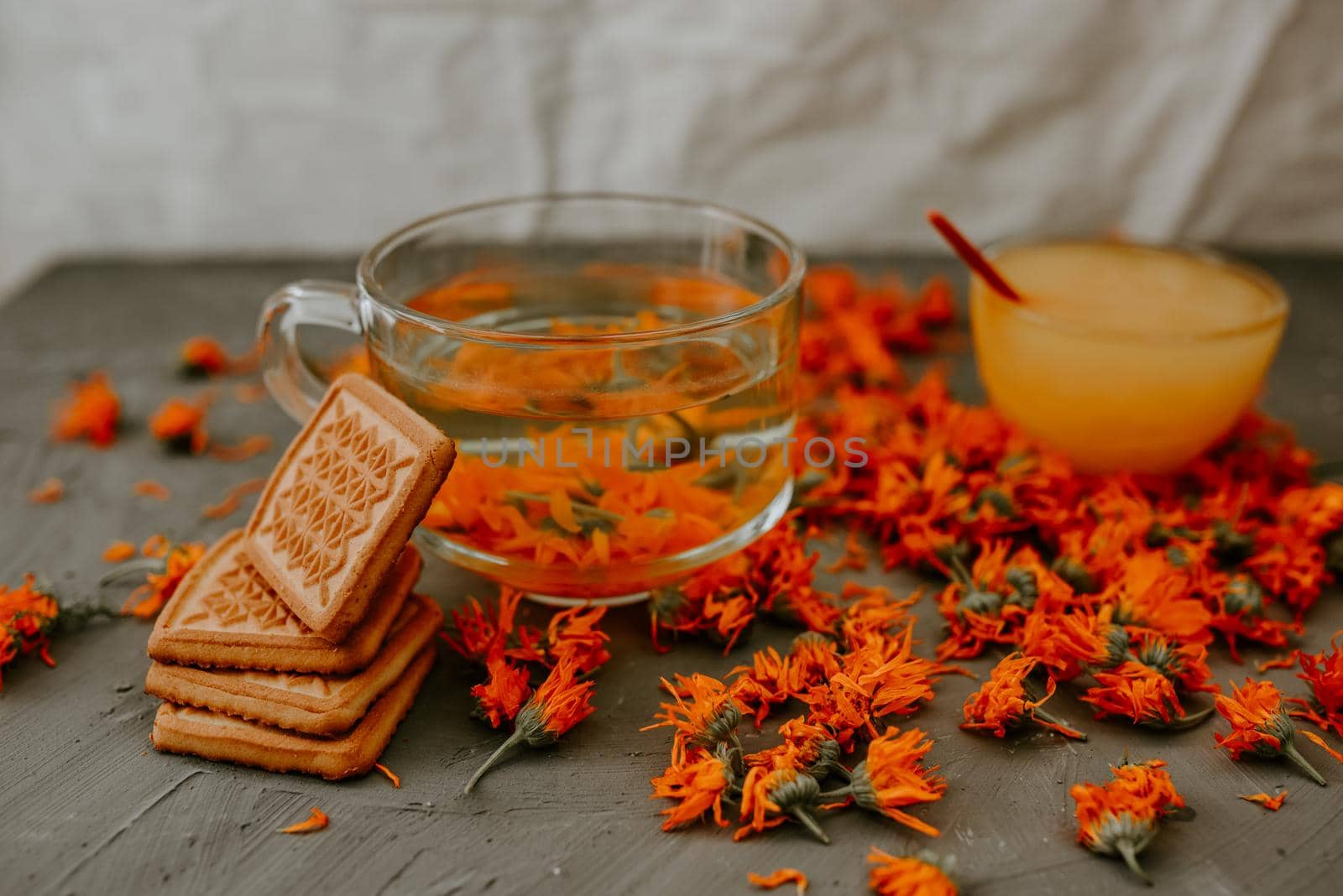 Tea with calendula flowers and biscuits. Transparent glass cup and saucer. Medicinal herbal dried plants marigold, orange calendula. Neutral white and gray background.