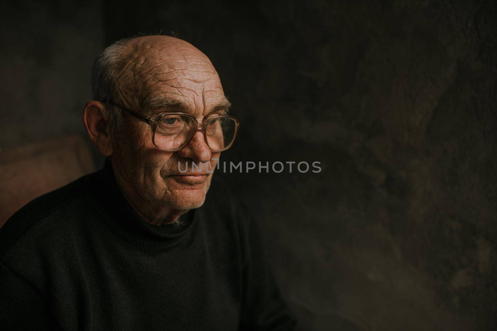 Pensive old man in glasses with gray hair looks away. wrinkles. wisdom. against a dark gray texture wall. bald head. in a knitted sweater. Portrait.