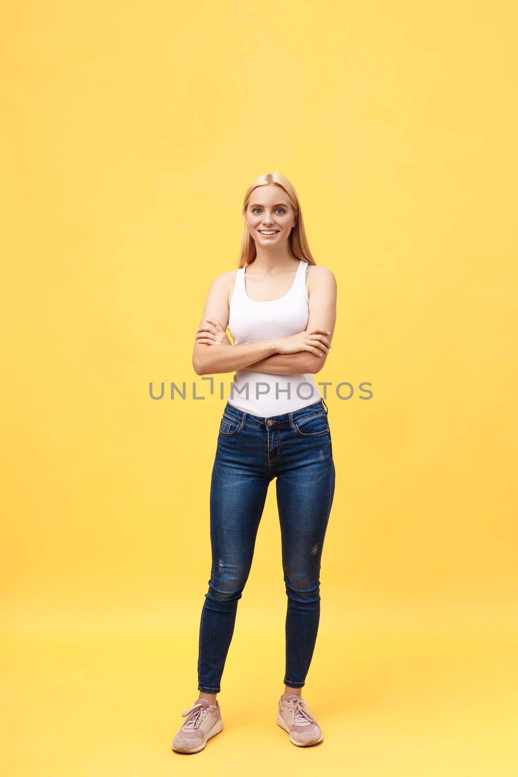 Full length photo of beautiful smiling woman standing with crossed hands, looking at camera, isolated on yellow background