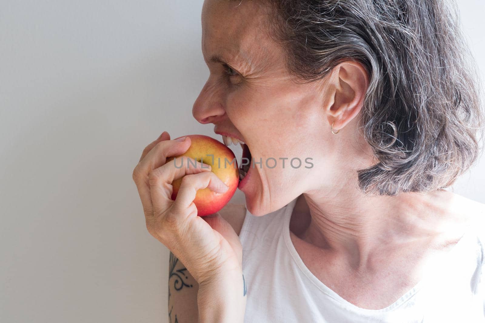 Profile of middle aged woman with grey hair dramatically biting into red apple (selective focus)