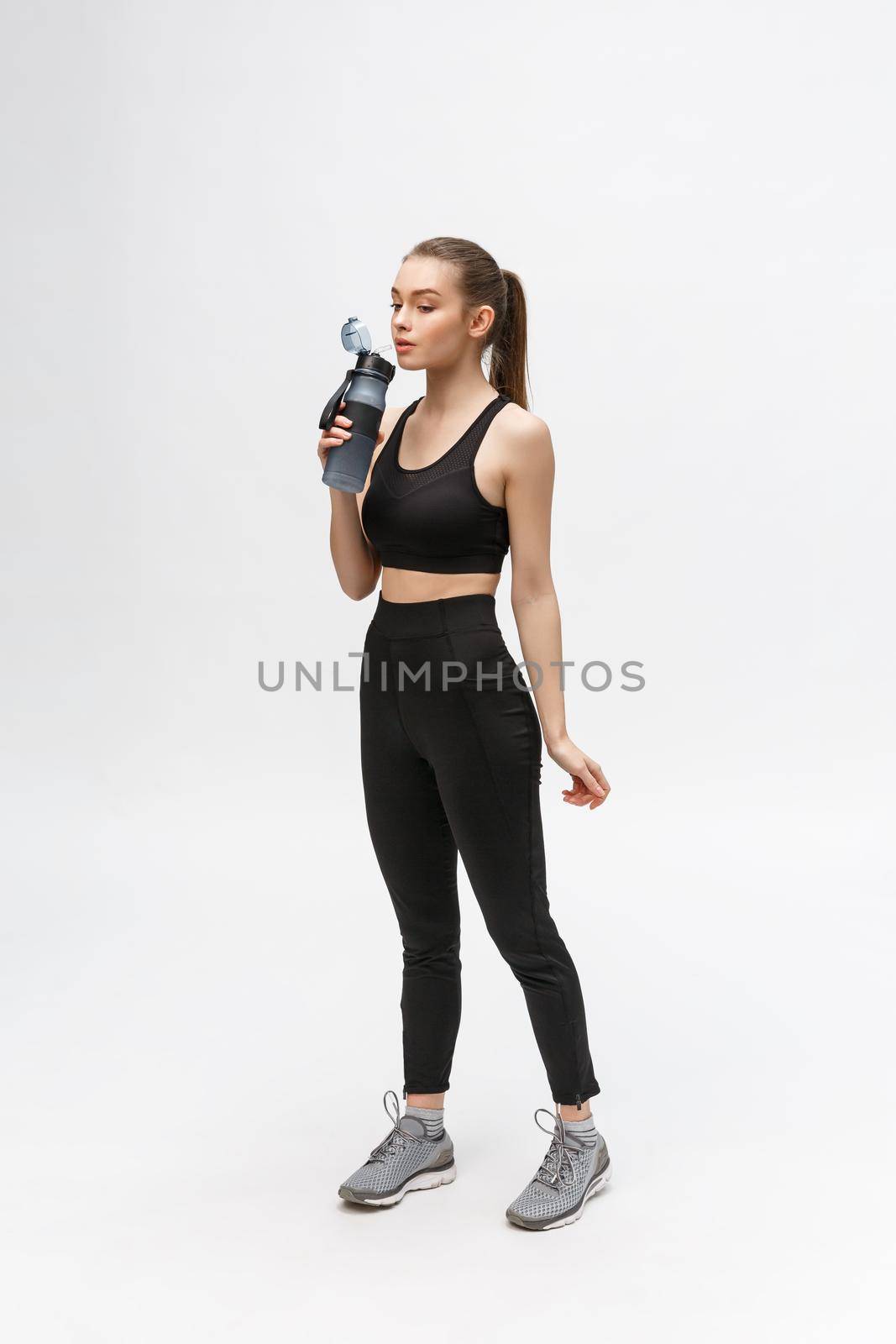 Side view full length portrait of a young healthy sports woman holding a water bottle isolated on a white background by Benzoix