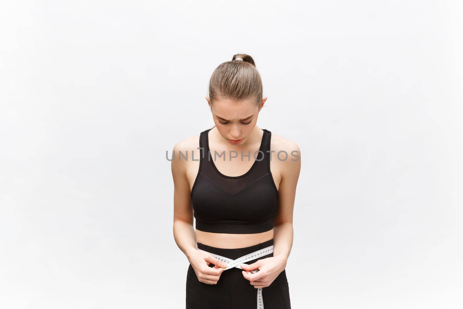 Young woman with excess weight in sporty top sadly looking on result of measuring waist over white background.