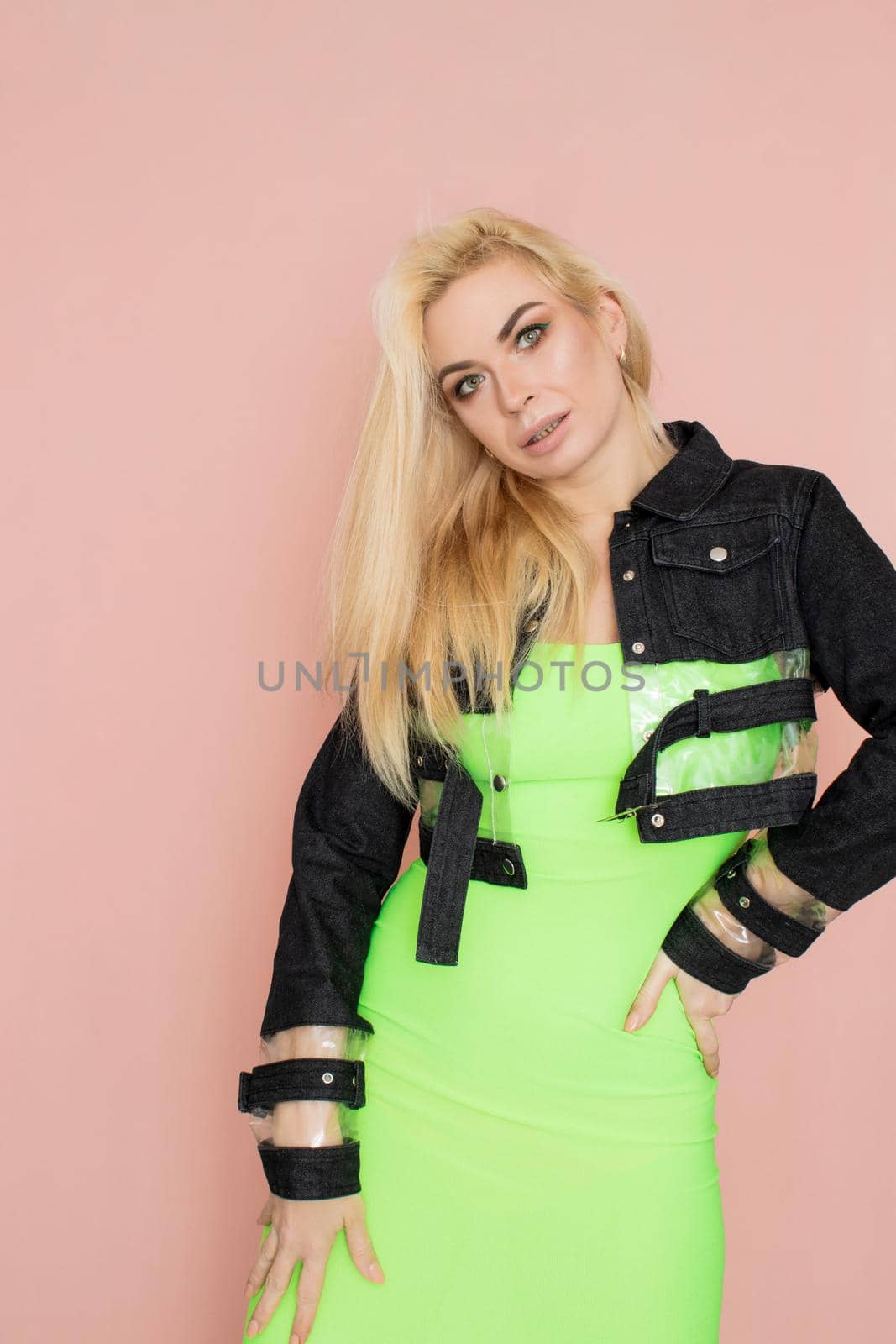 Stylish caucasian adult blonde woman wears fashionable jeans jacket and green dress, isolated over pink studio wall.
