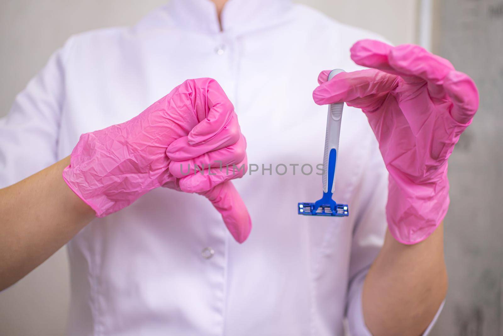 A close-up of a woman in a medical gown is hands in pink gloves holding a razor. Thumb down indignantly shows the depilation master