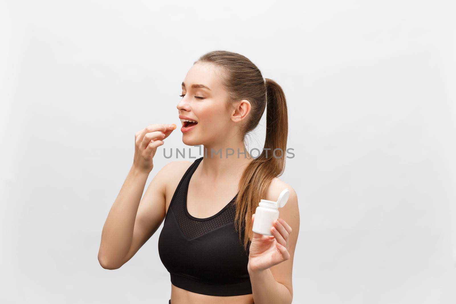 Smiling blonde sport woman holding bottle of medicine on white background by Benzoix