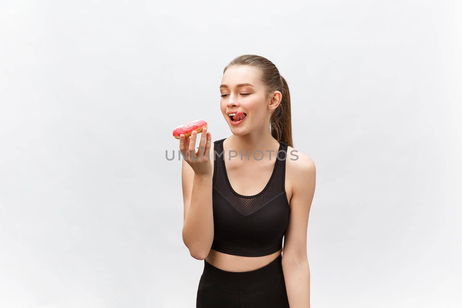 Beautiful Fitness model wearing a black fitness outfit holding a junk food donut. Isolated over grey background. by Benzoix