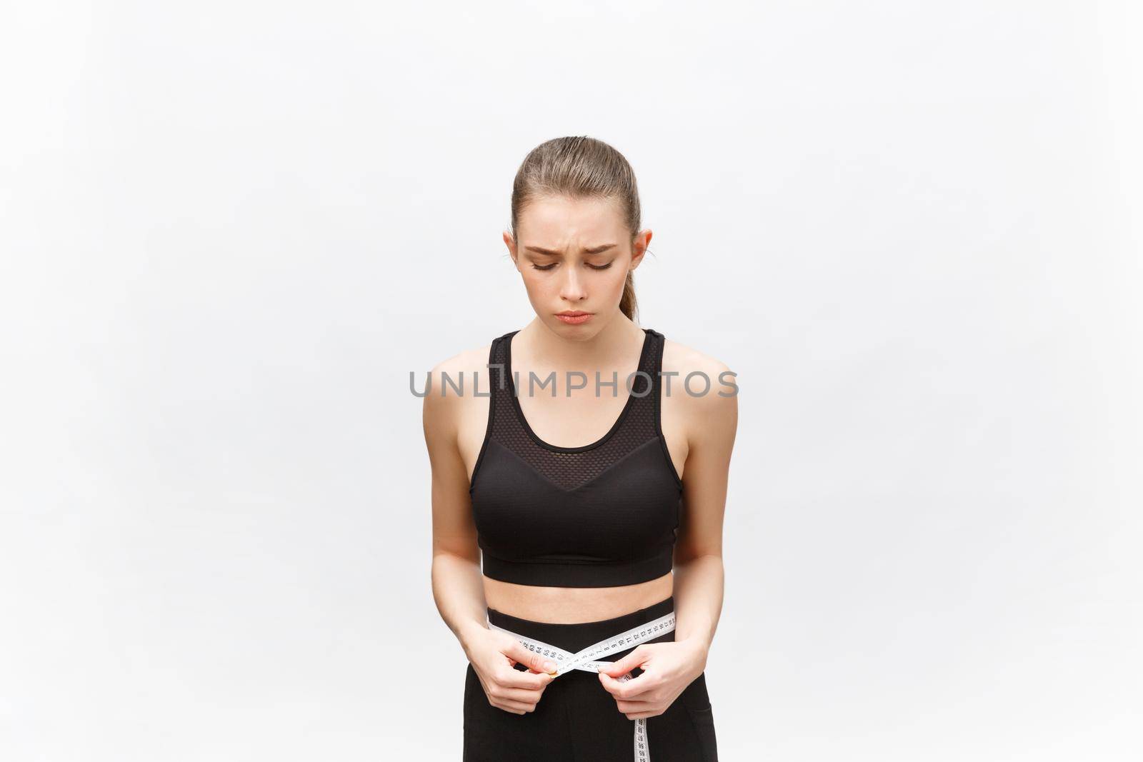 Young woman with excess weight in sporty top sadly looking on result of measuring waist over white background by Benzoix