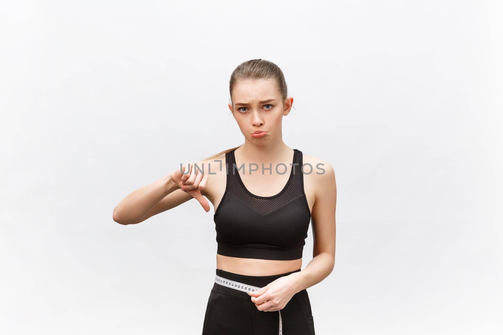 Young woman with excess weight in sporty top sadly looking on result of measuring waist over white background by Benzoix