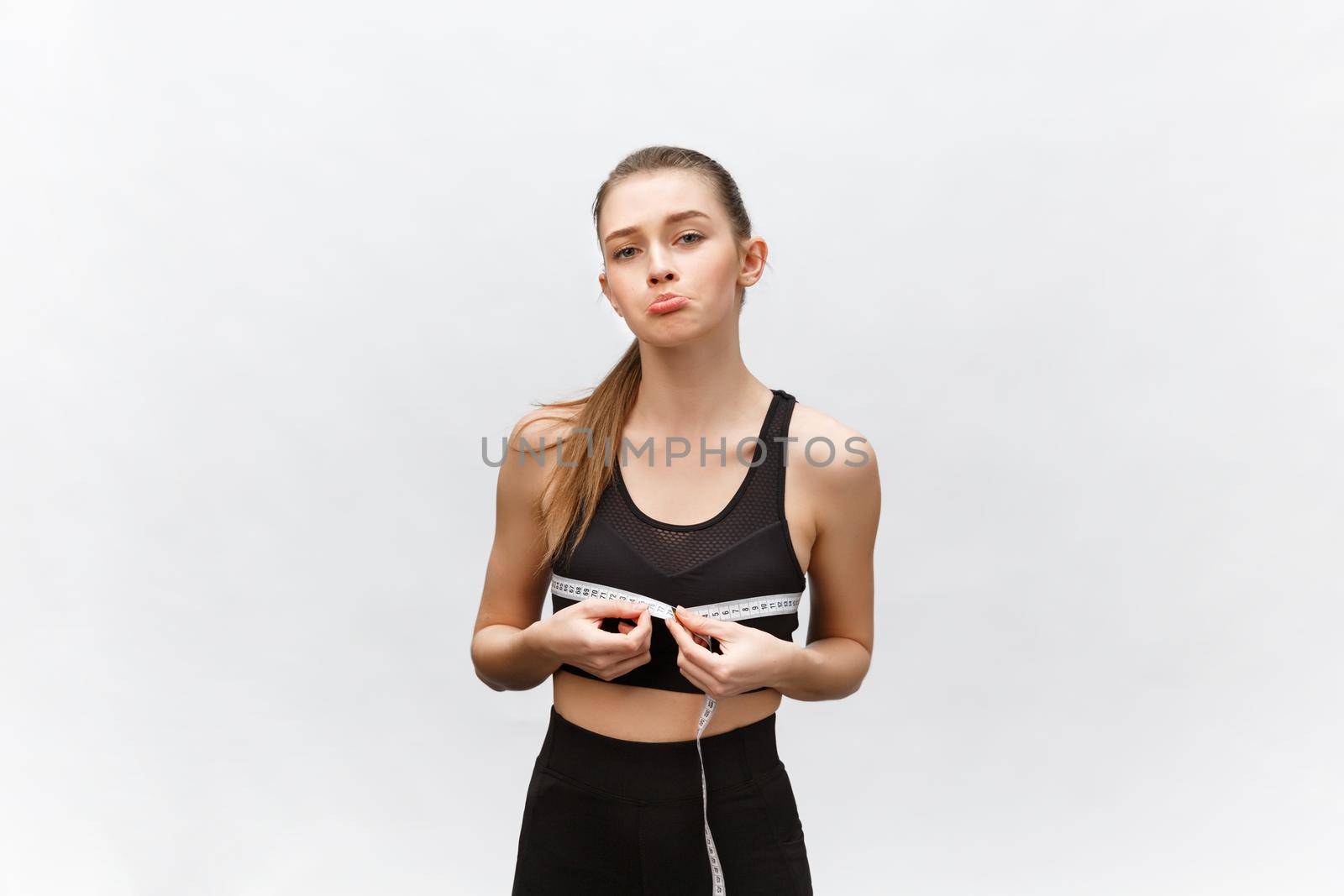 Young woman with excess weight in sporty top sadly looking on result of measuring waist over white background by Benzoix