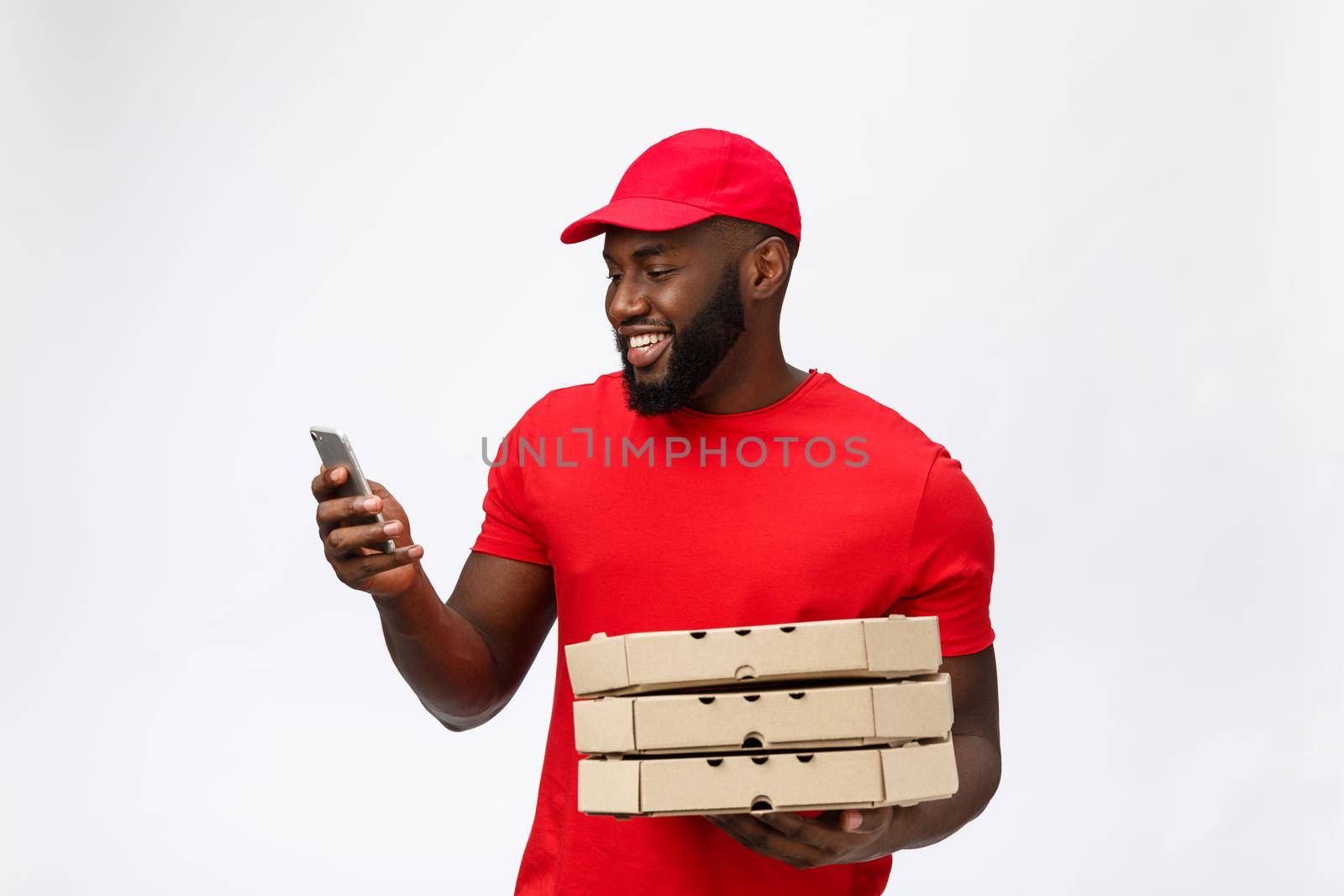 Delivery Concept: Handsome african pizza delivery man talking to mobile with happy facial expression. Isolated over grey background. by Benzoix