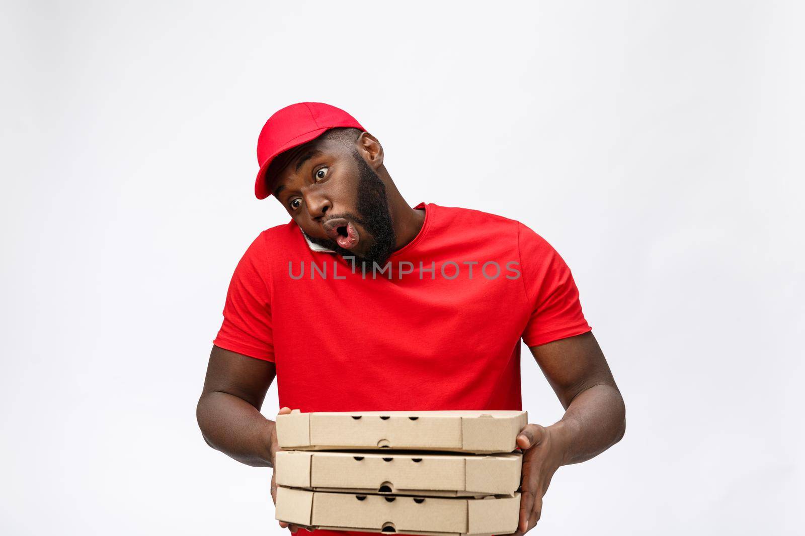 Delivery Concept: Handsome african pizza delivery man talking to mobile with shocking facial expression. Isolated over grey background. by Benzoix