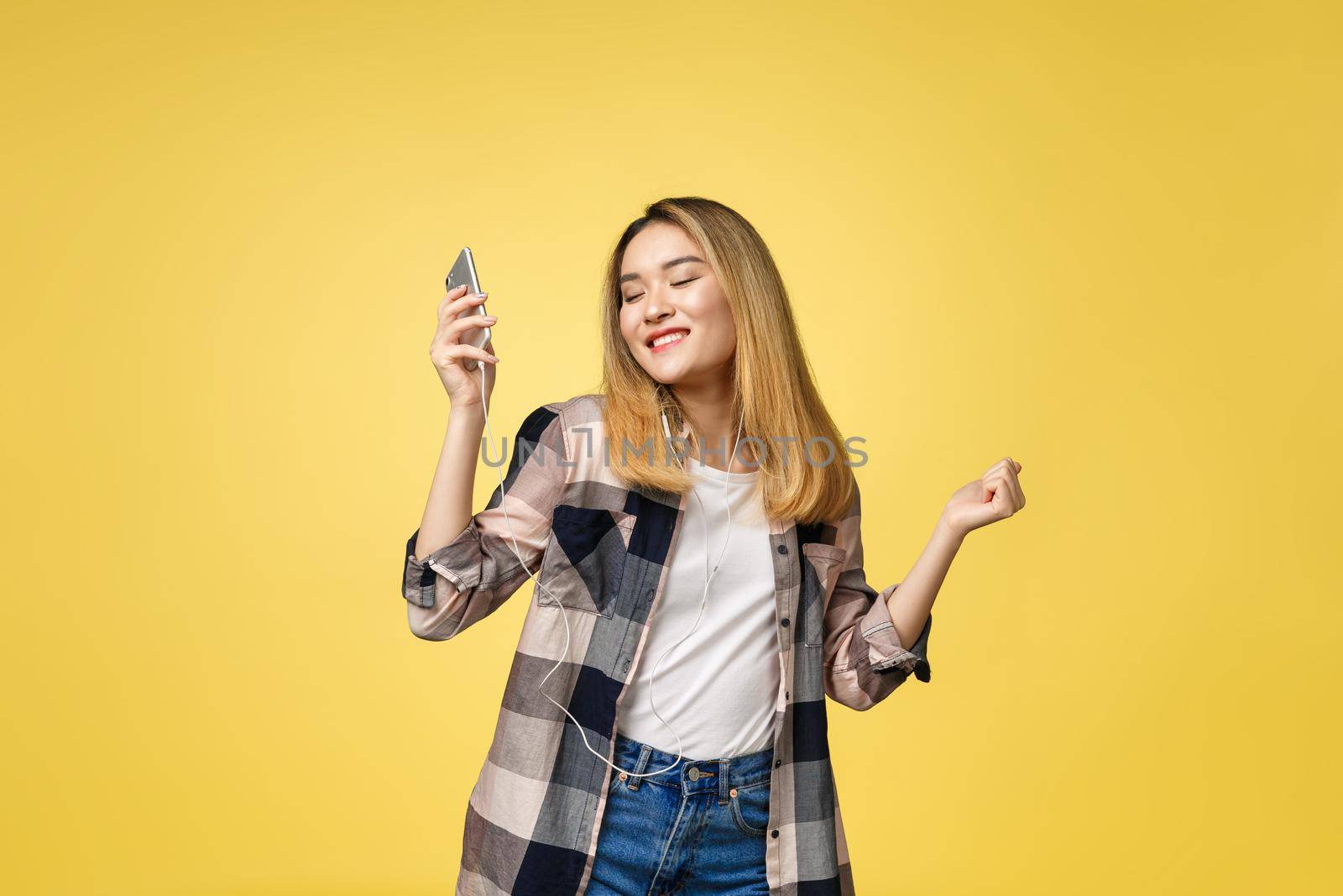 Fashion smiling asian woman listening to music in earphones over yellow background. by Benzoix