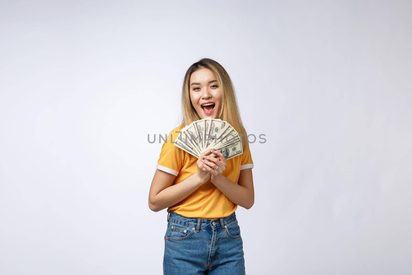 Asian woman holding cash notes isolated in white background. Young asian woman in white t-shirt in winning surprise, holding dollar note. Young rich hipster concept. by Benzoix