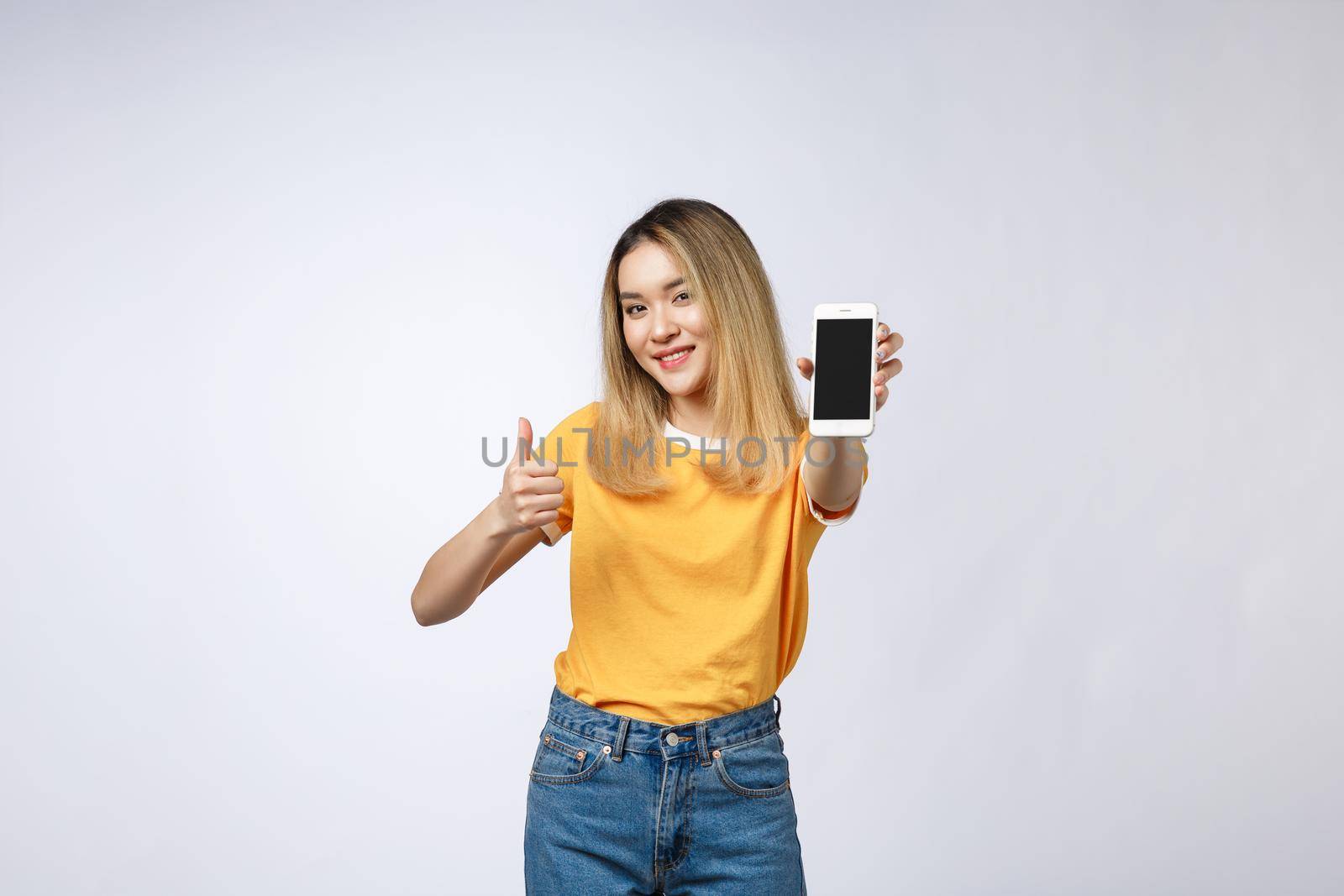 Young Asian woman wearing in yellow shirt is showing thumb up sign on white background, holding mobile phone, smiling
