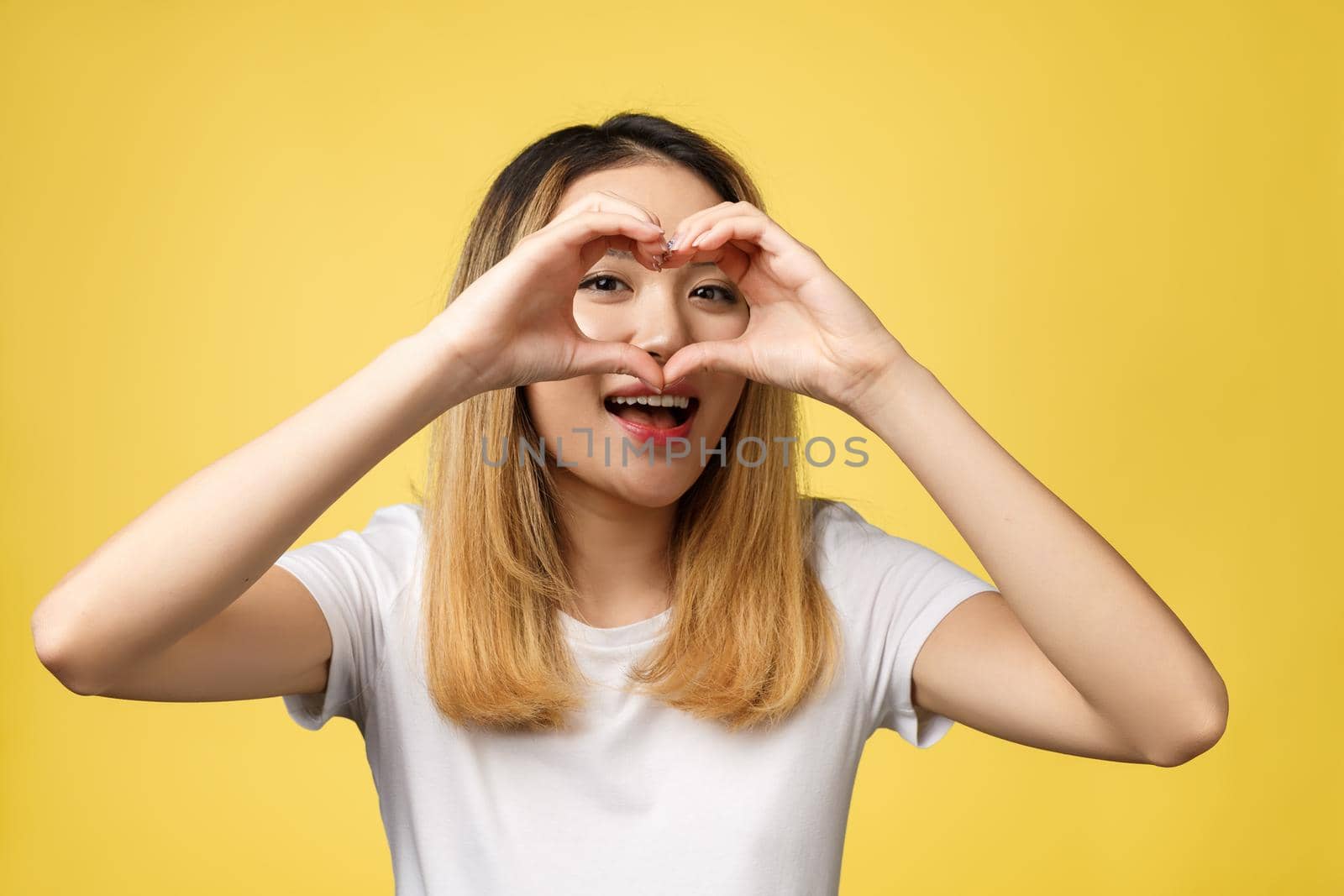 Young Asian woman show heart hand sign isolated on yellow background