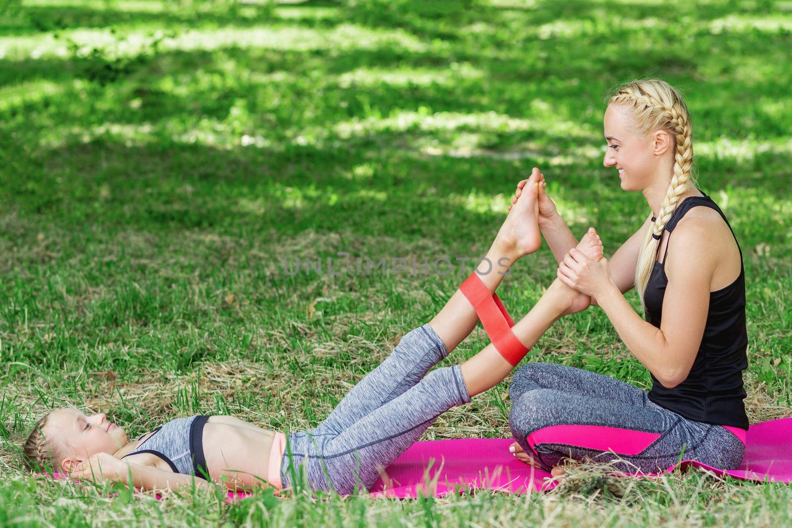 Young woman is training legs of little girl by fitness gum on mat in the park.