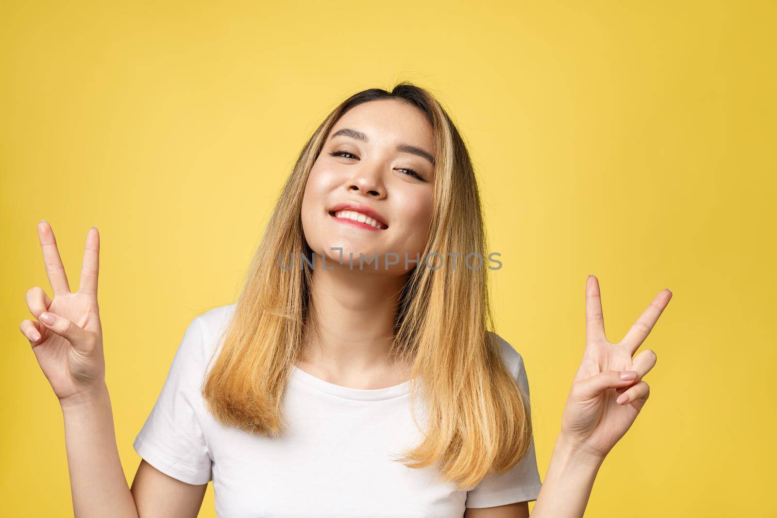 Pleased asian woman in t-shirt showing peace gestures and looking at the camera over yellow background by Benzoix