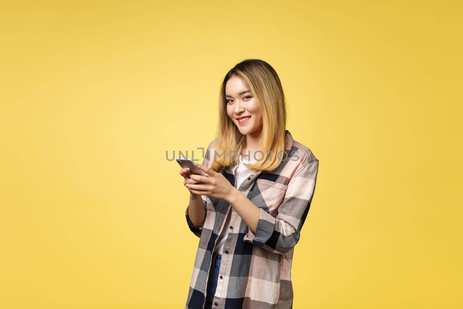 Image of satisfied asian woman with long hair smiling and texting on cell phone holding in hand isolated over yellow background by Benzoix