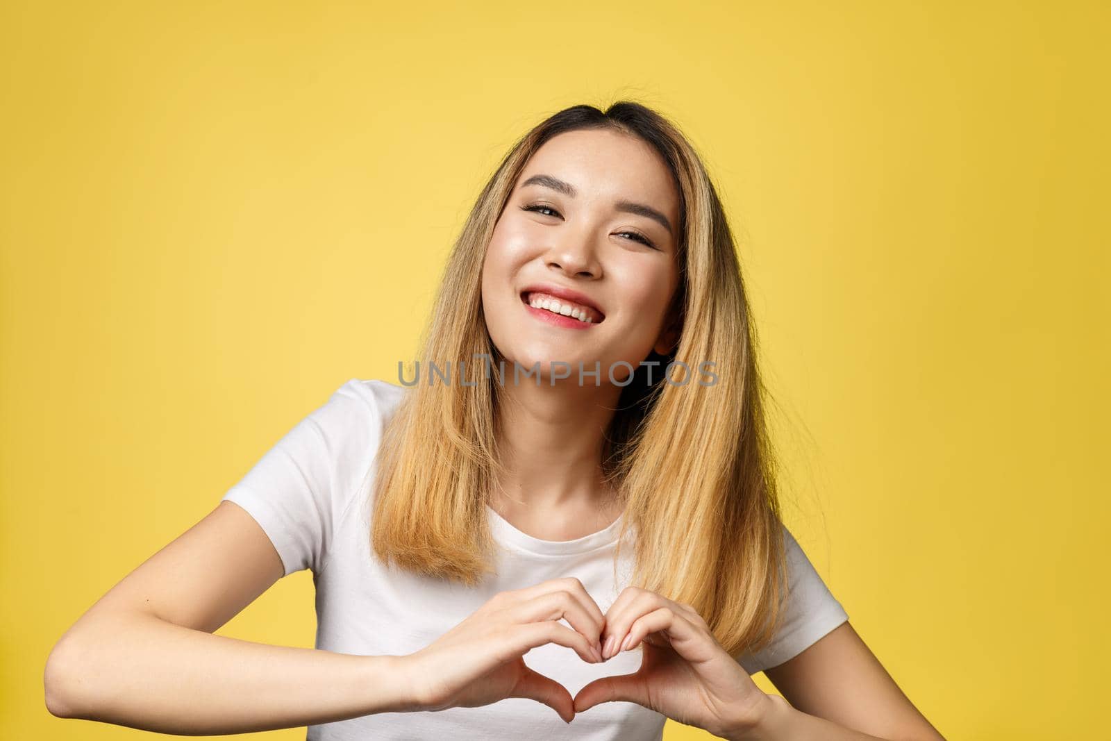 Young Asian woman show heart hand sign isolated on yellow background. by Benzoix