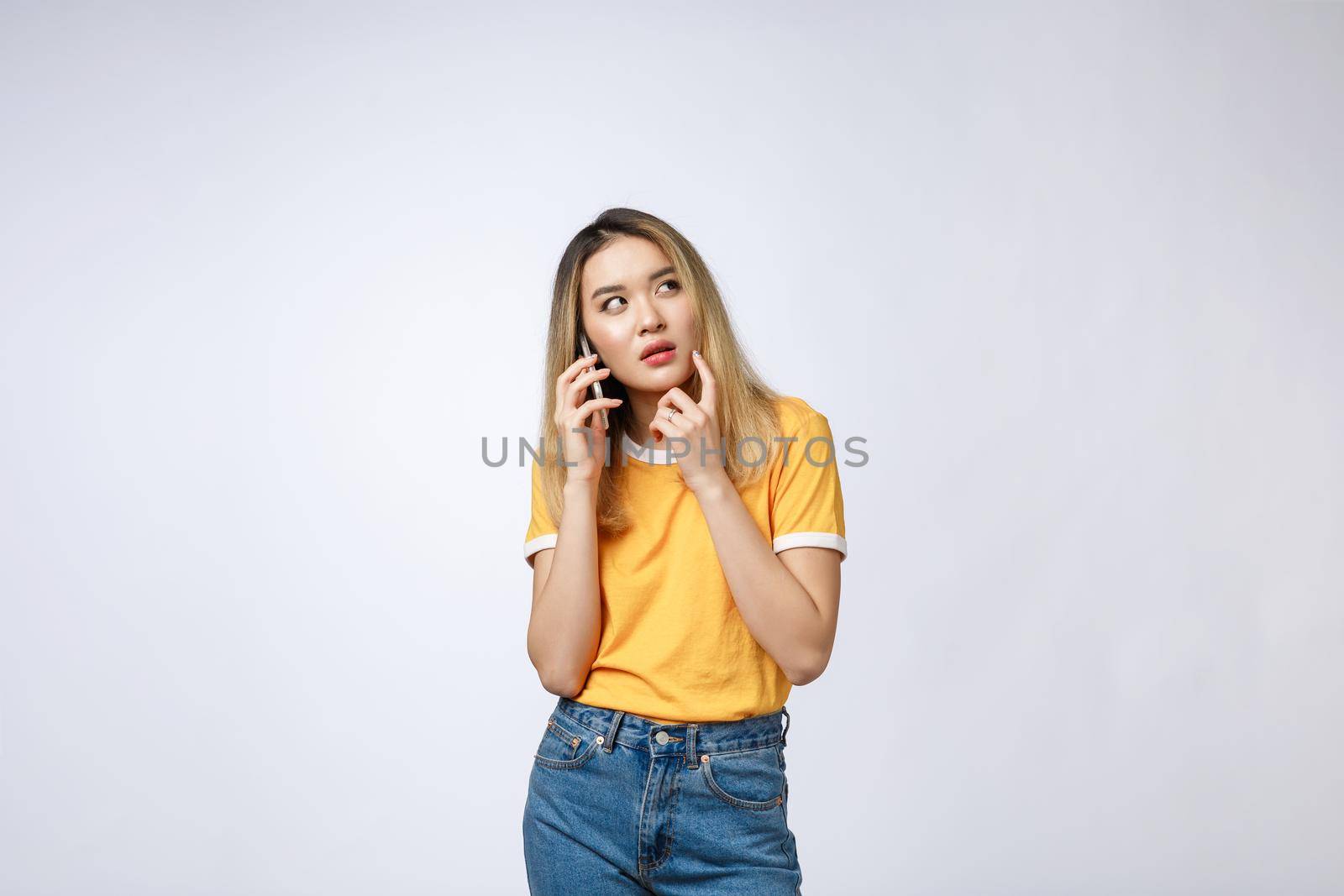 Portrait of a pensive young asian woman holding mobile phone and looking away thinking isolated over white background.