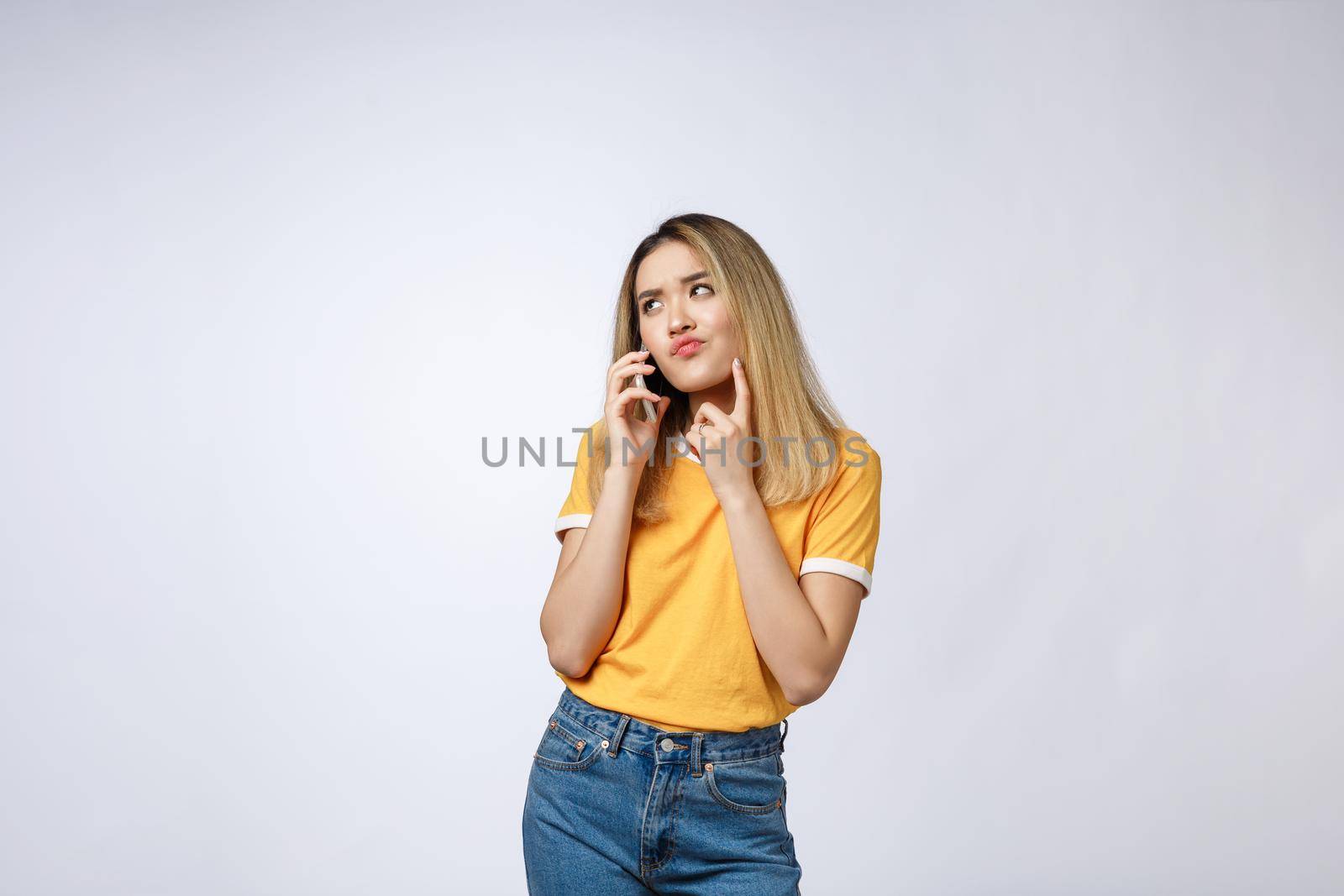 Portrait of a pensive young asian woman holding mobile phone and looking away thinking isolated over white background.