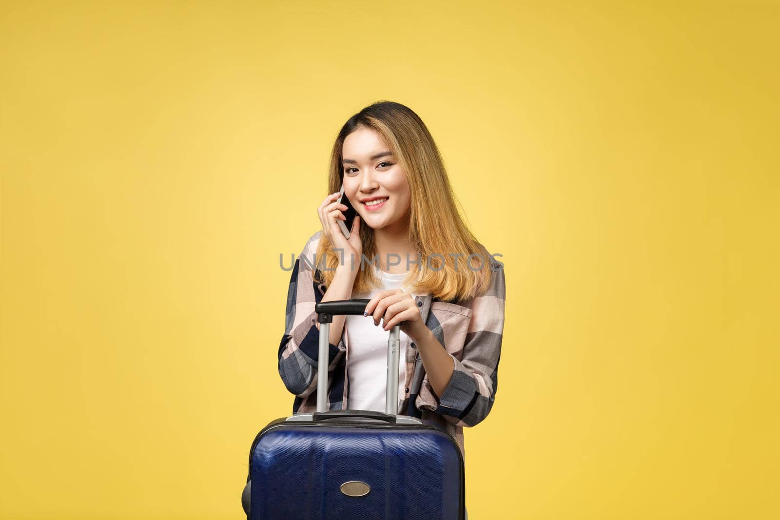 Portrait of happy asian female traveler with suitcase and looking at cellphone against isolated yellow background by Benzoix