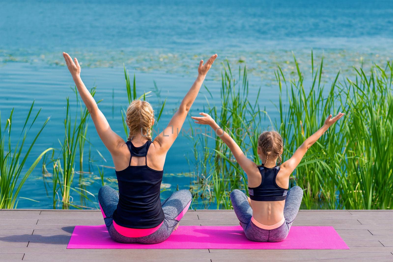 Mother and daughter doing yoga at the shore of the lake in summer.