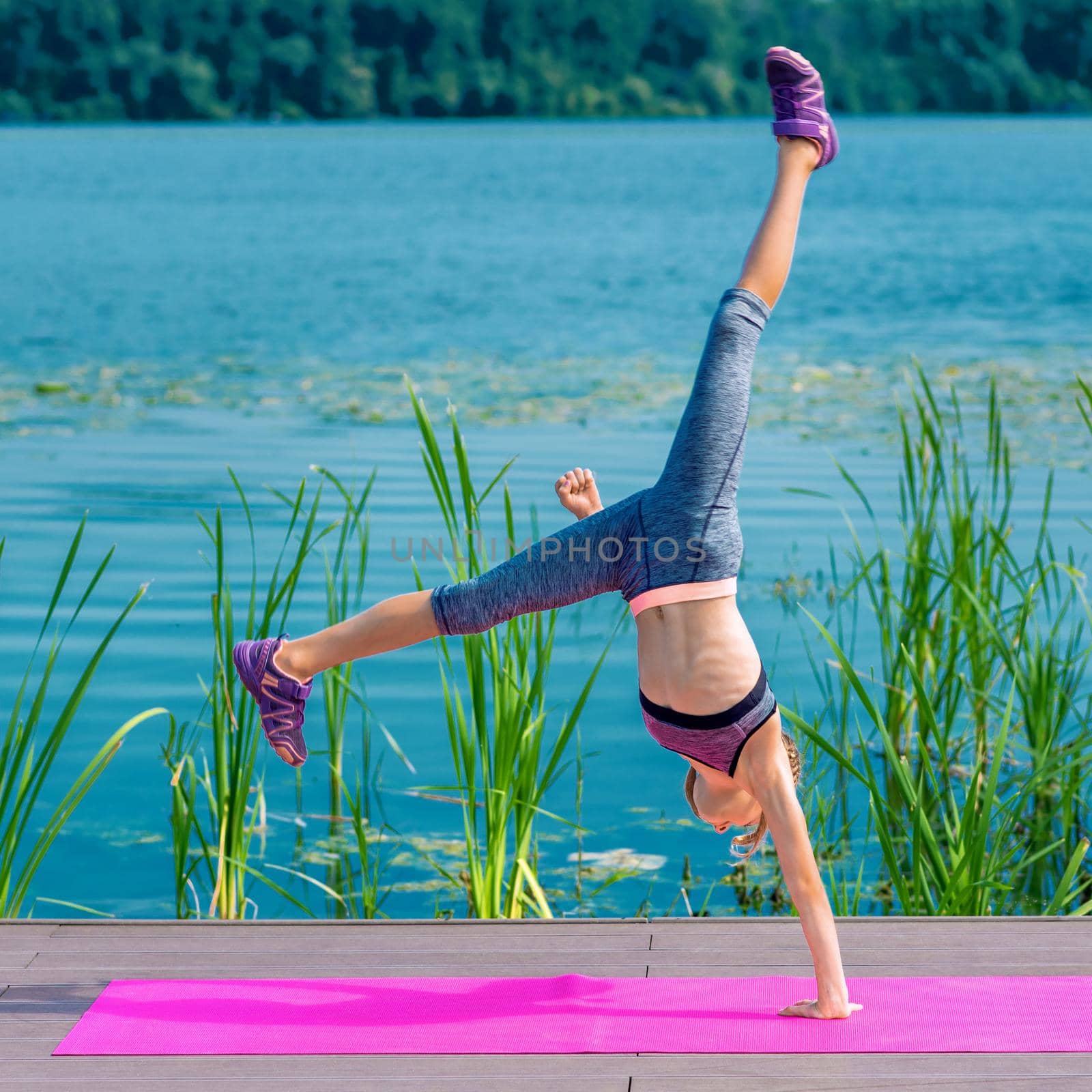 Fit sporty little girl in sportswear doing cartwheel exercise on the roll mat by the lake.