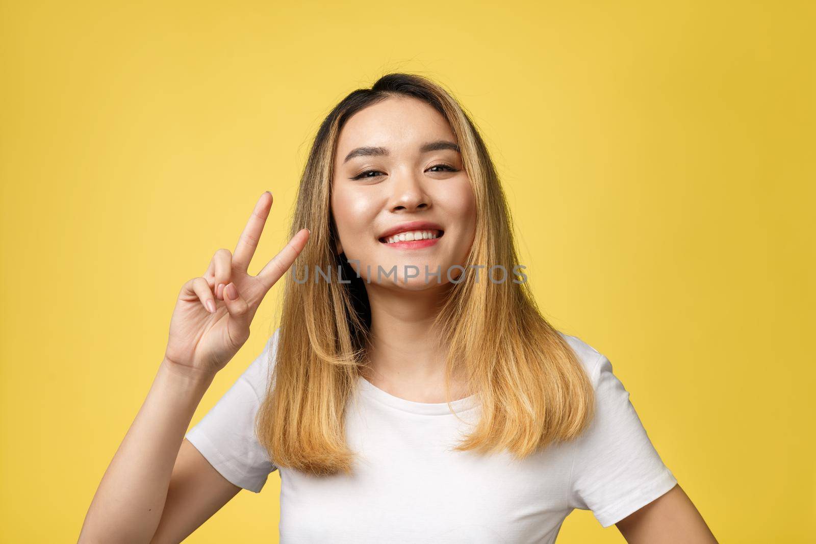 Pleased asian woman in t-shirt showing peace gestures and looking at the camera over yellow background.