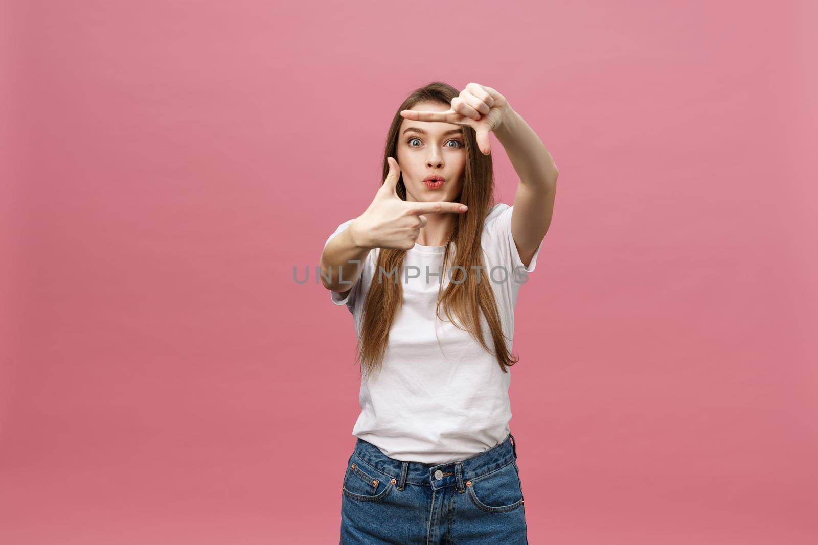 Cheerful young woman keeping mouth wide open, looking surprised, making hands photo frame gesture isolated on bright pink background