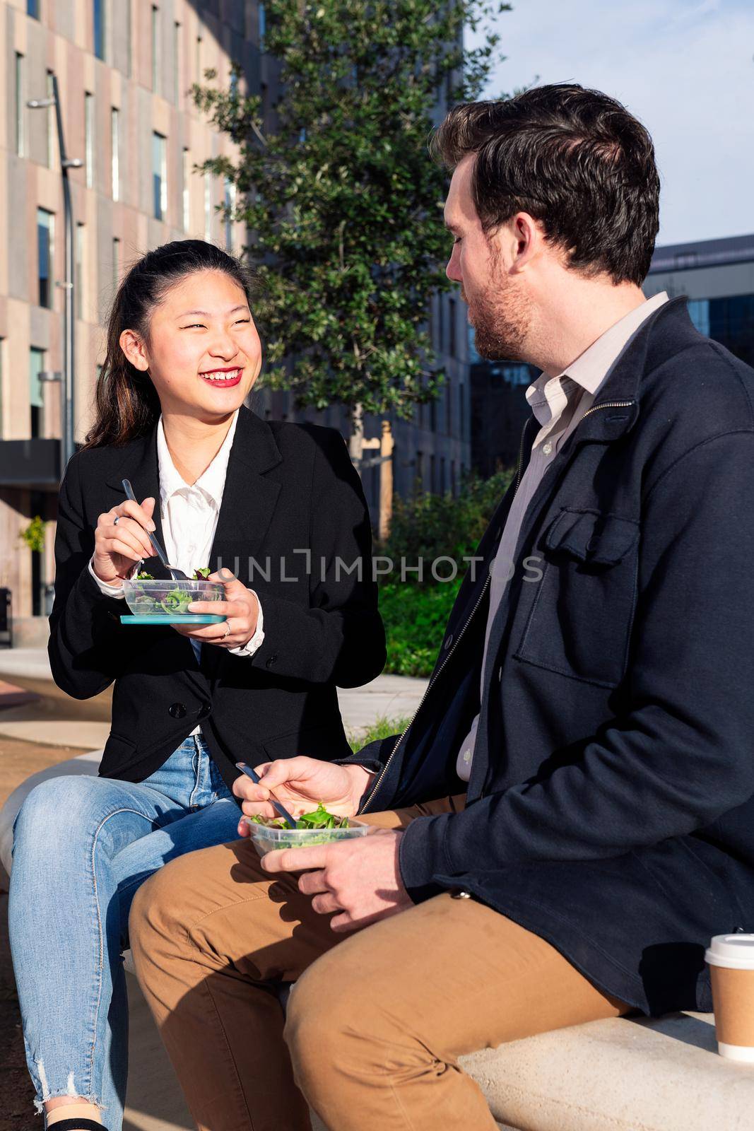 asian woman smiling and eating a salad with her caucasian partner in a park next to the office, concept of healthy fast food at work