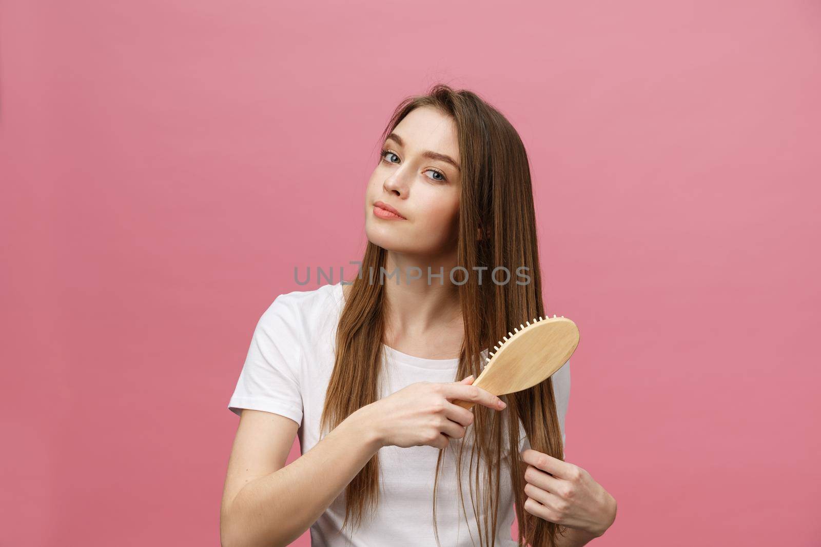 Smiling young woman combing hair and looking away isolated on pink.