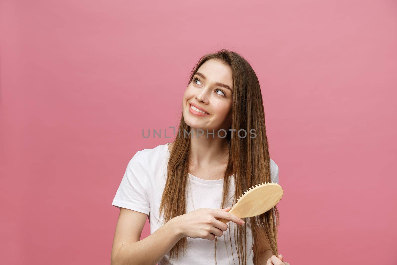 Smiling young woman combing hair and looking away isolated on pink by Benzoix