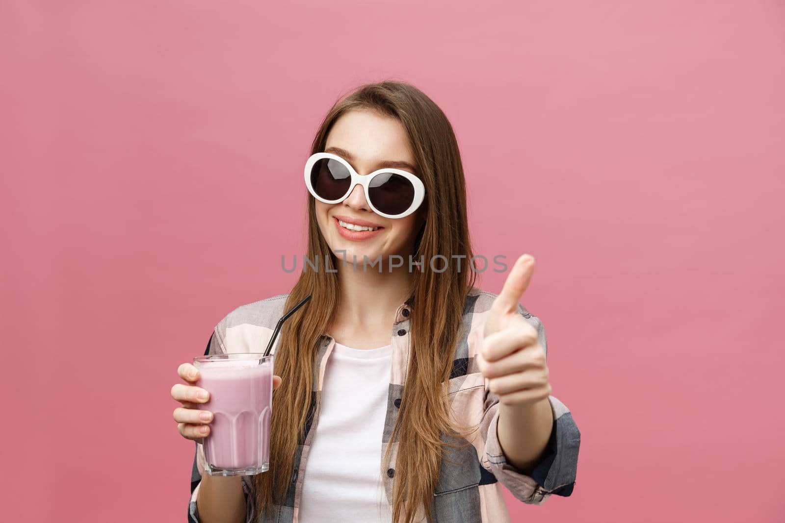 Portrait of a cute casual girl drinking orange juice from a glass and looking at camera isolated over pink background.