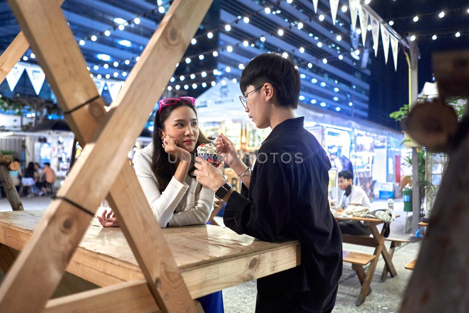 Non binary person feeding ice cream to her partner at urban night fair in Bangkok, Thailand