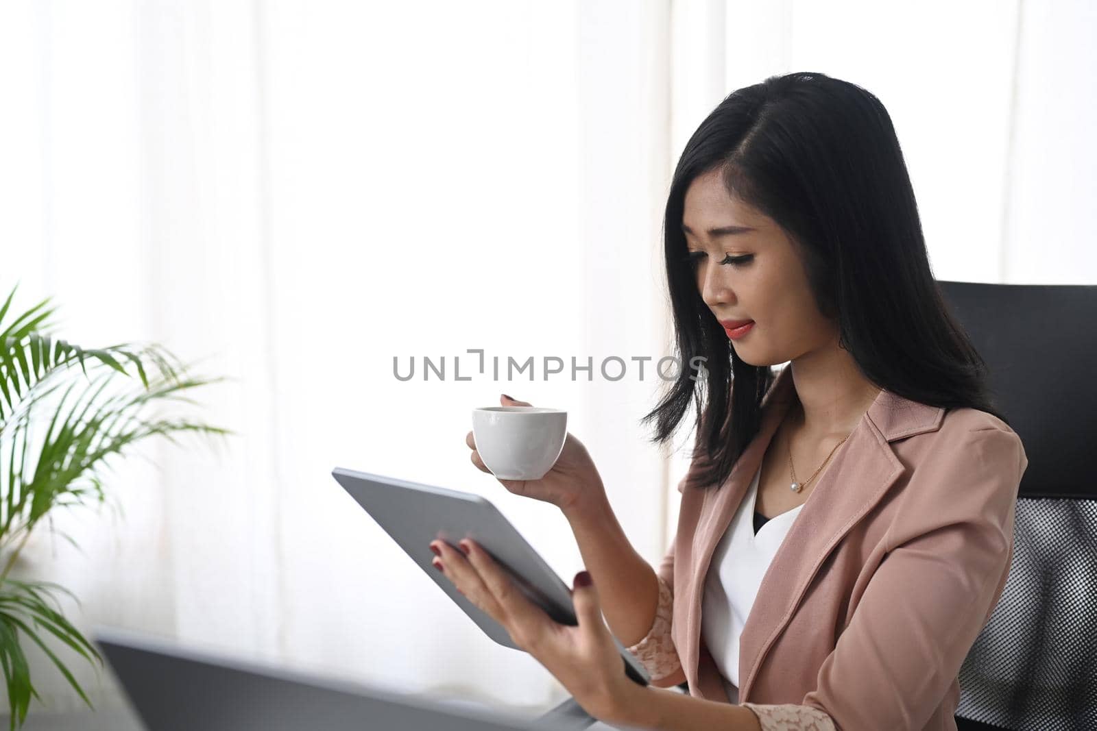 Young businesswoman drinking coffee and reading email on her digital tablet.
