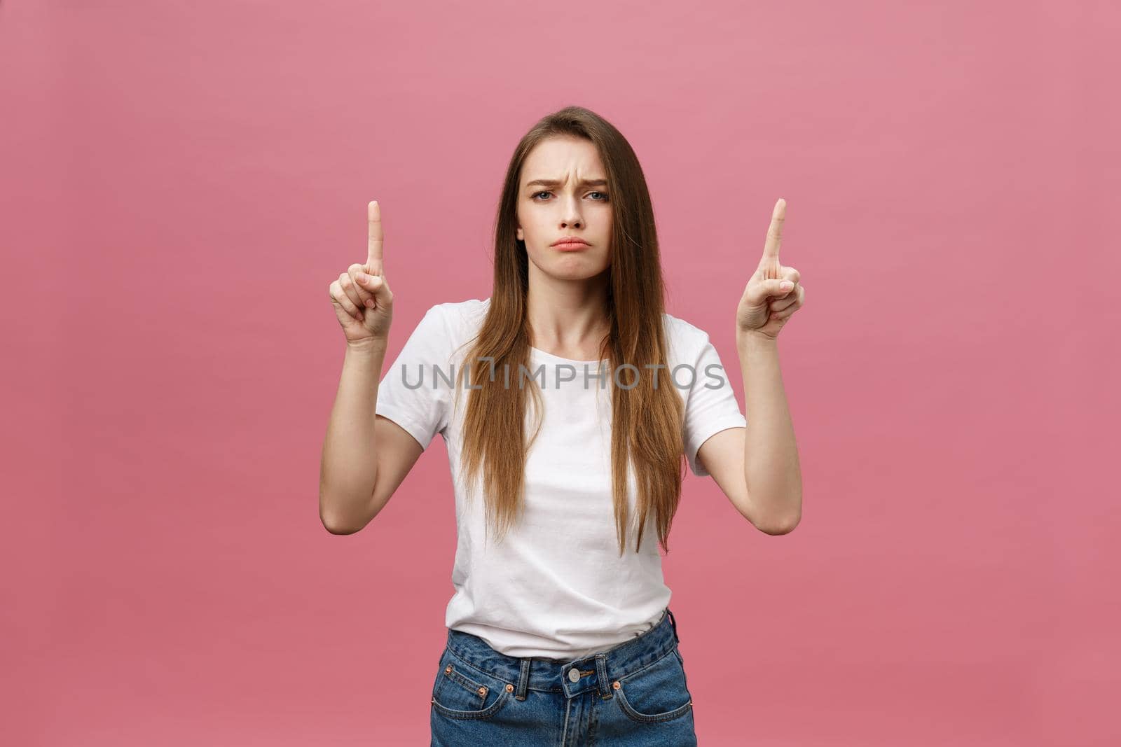 Closeup of serious strict young woman wears white shirt looks stressed and pointing up with finger isolated over pink background.