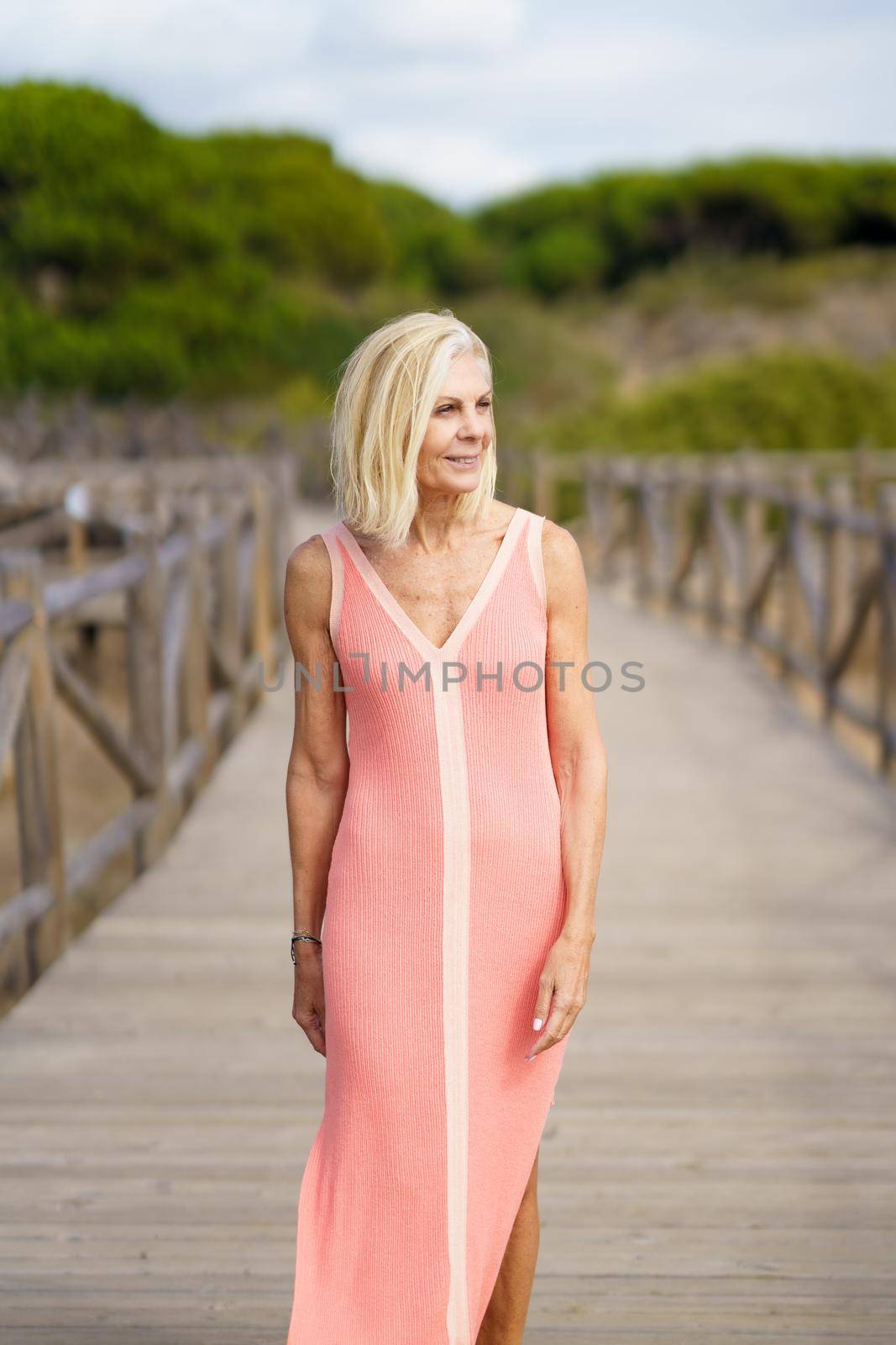 Beautiful mature woman walking along a wooden path near the beach., wearing a nice orange dress. Elderly female enjoying her retirement at a seaside retreat.