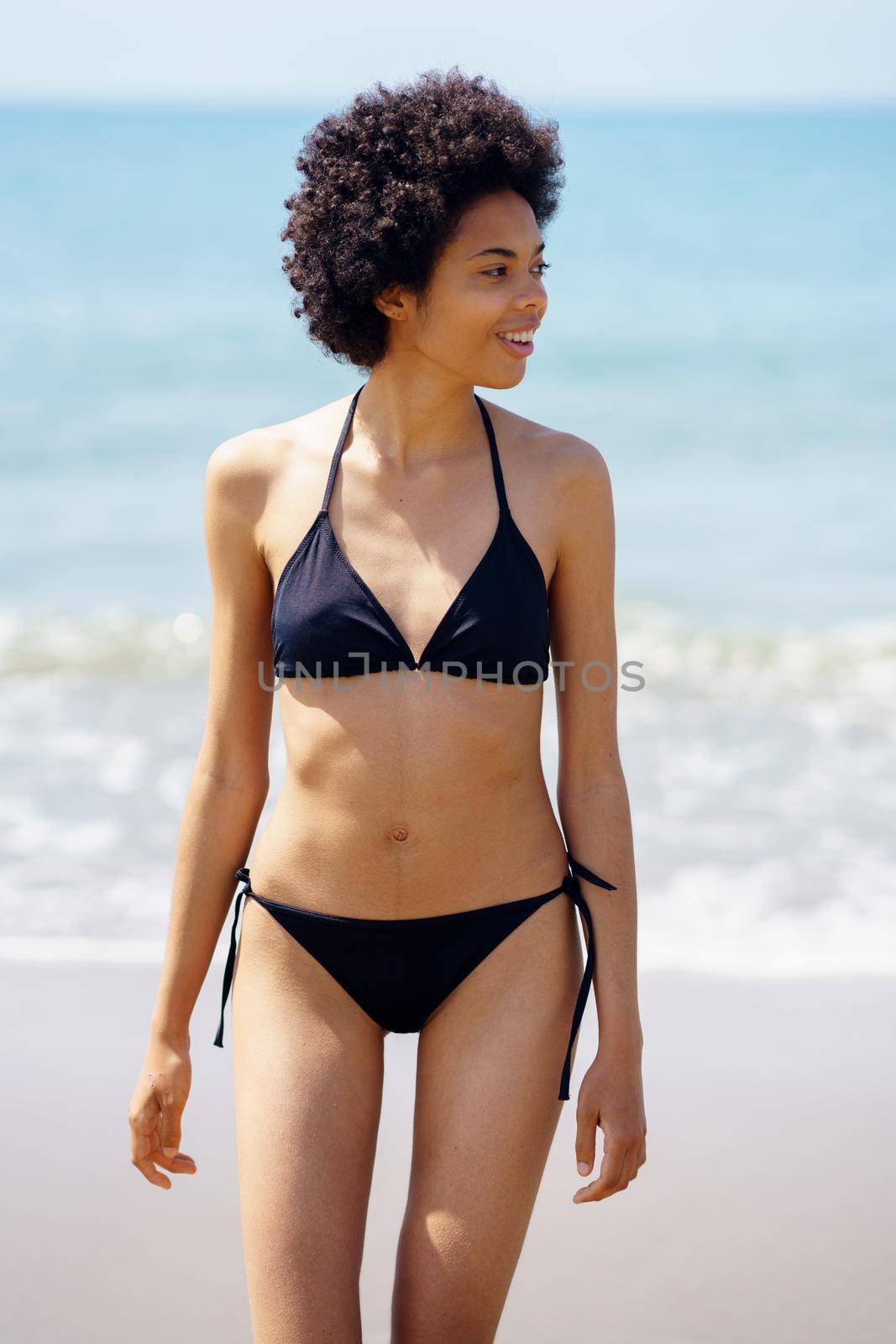 Positive black woman in bikini walking on the beach smiling. Girl with afro hairstyle coming out of the water at the beach.