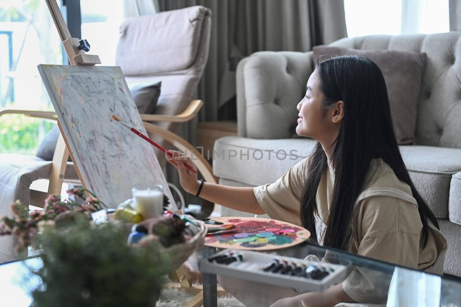 Cute little girl sitting on floor in living room and painting on canvas with watercolor.