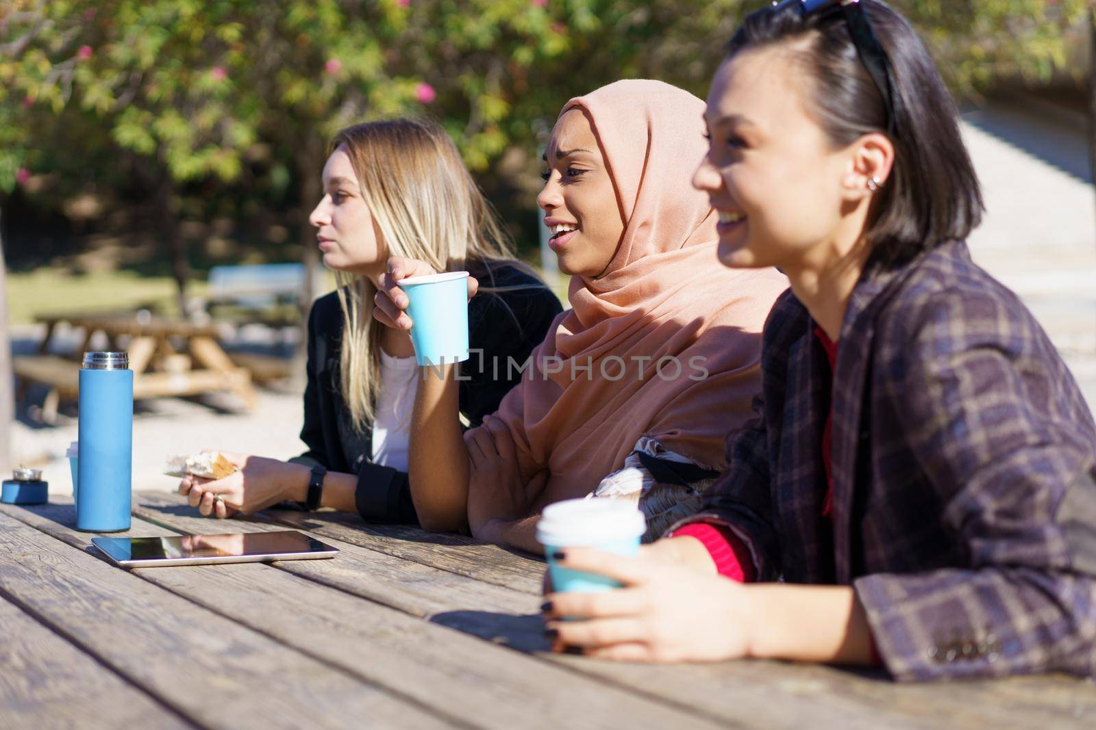 Positive young multiethnic female friends in casual clothes smiling while resting in outdoor cafe and drinking coffee from disposable cups