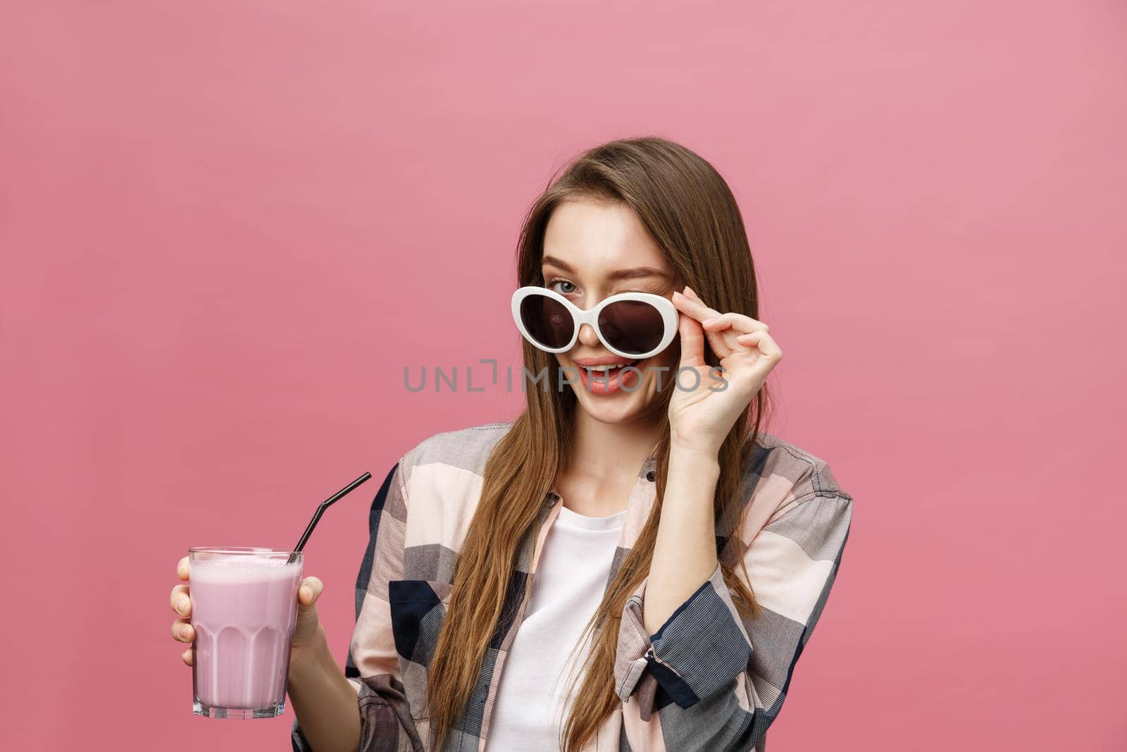 Portrait of a cute casual girl drinking orange juice from a glass and looking at camera isolated over pink background.