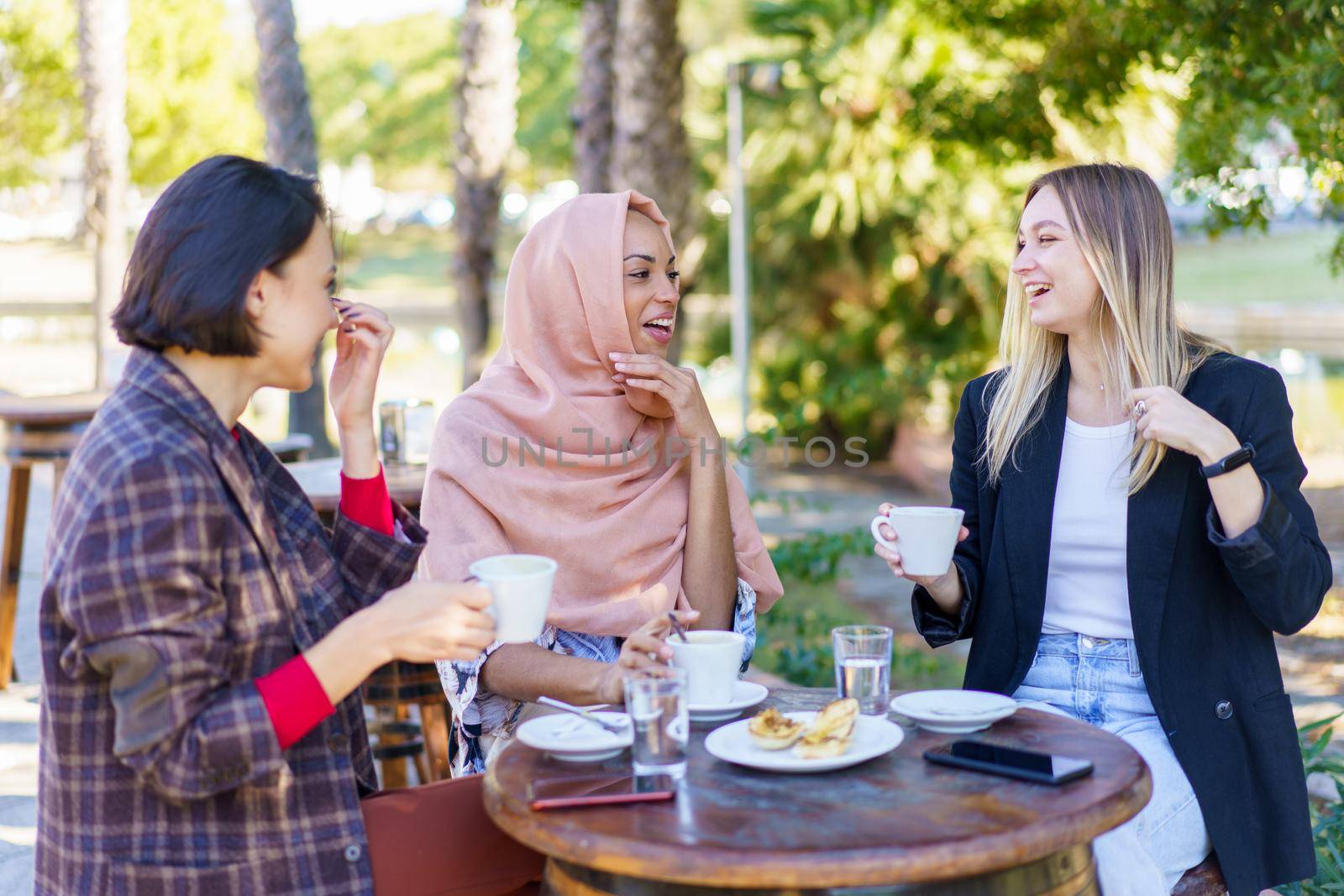 Cheerful diverse female friends looking at each other while drinking coffee at table on terrace of outdoor cafeteria on summer day
