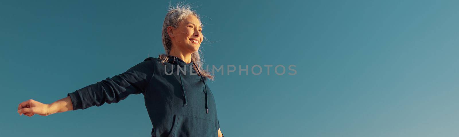 Smiling lady in a sportswear raising arms to the sides, training and looking ahead by the waterfront