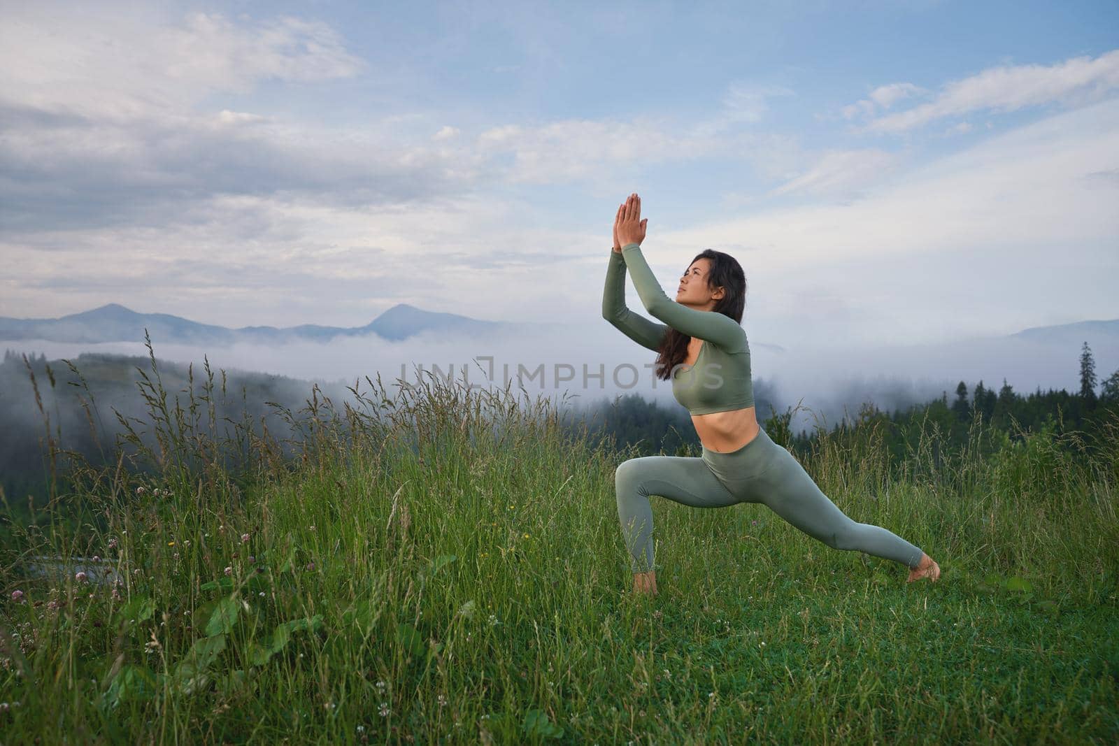 Young dark haired woman in sport outfit having yoga practice among green mountains. Concept of people, wellbeing and body care.
