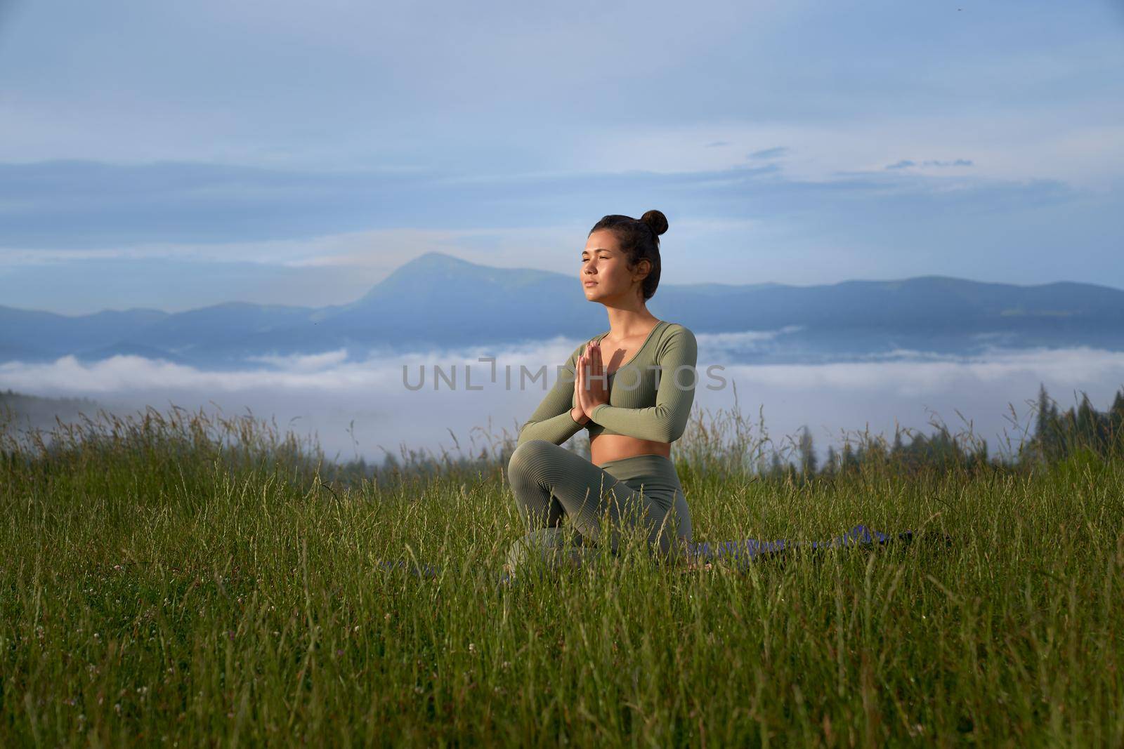 Sporty young woman sitting on yoga mat with namaste gesture and enjoying outdoors meditation. Concept of wellness, spirituality and harmony.