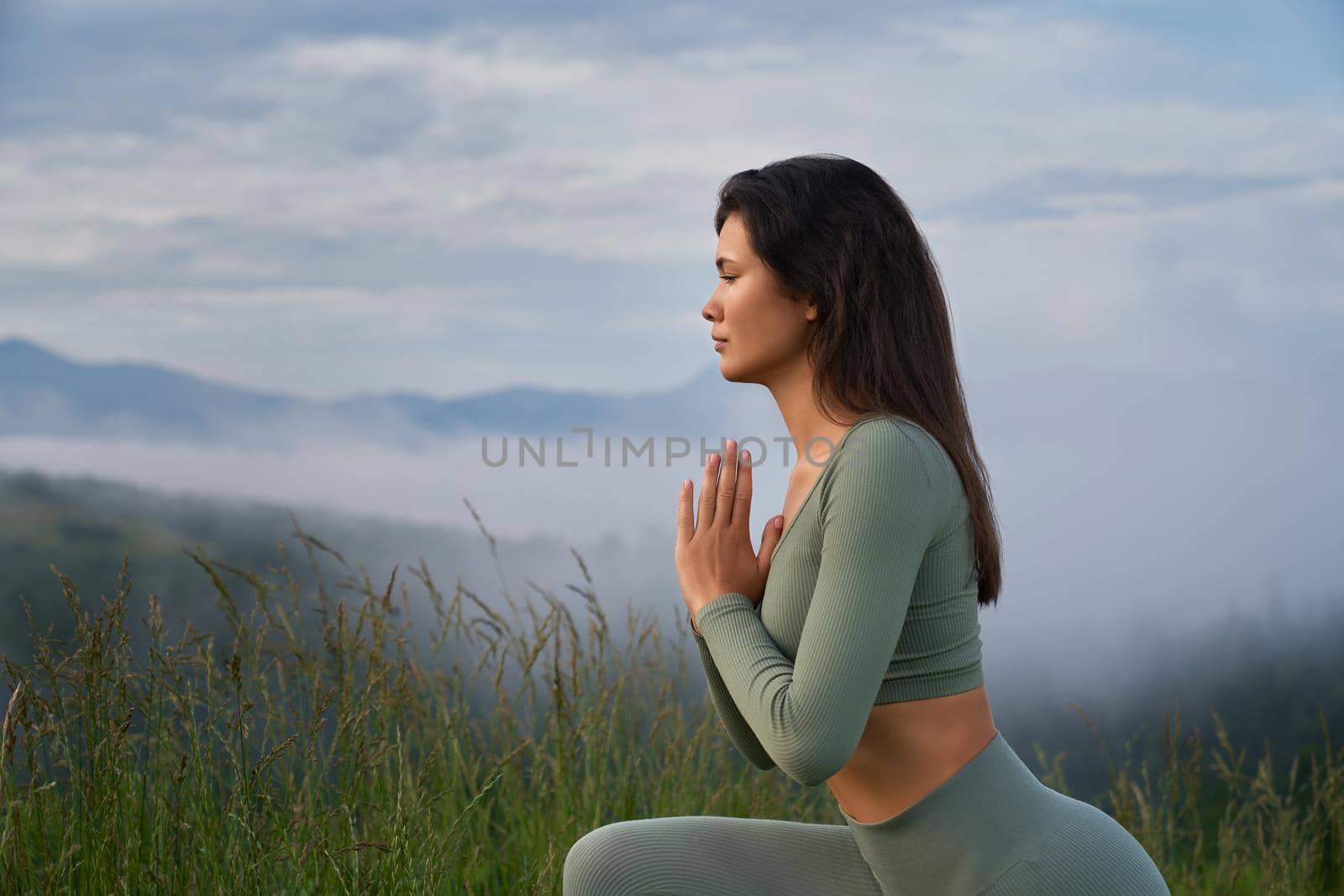 Side view of young woman with dark hair doing yoga exercises with namaste gesture among green summer mountains. Morning training on fresh air.