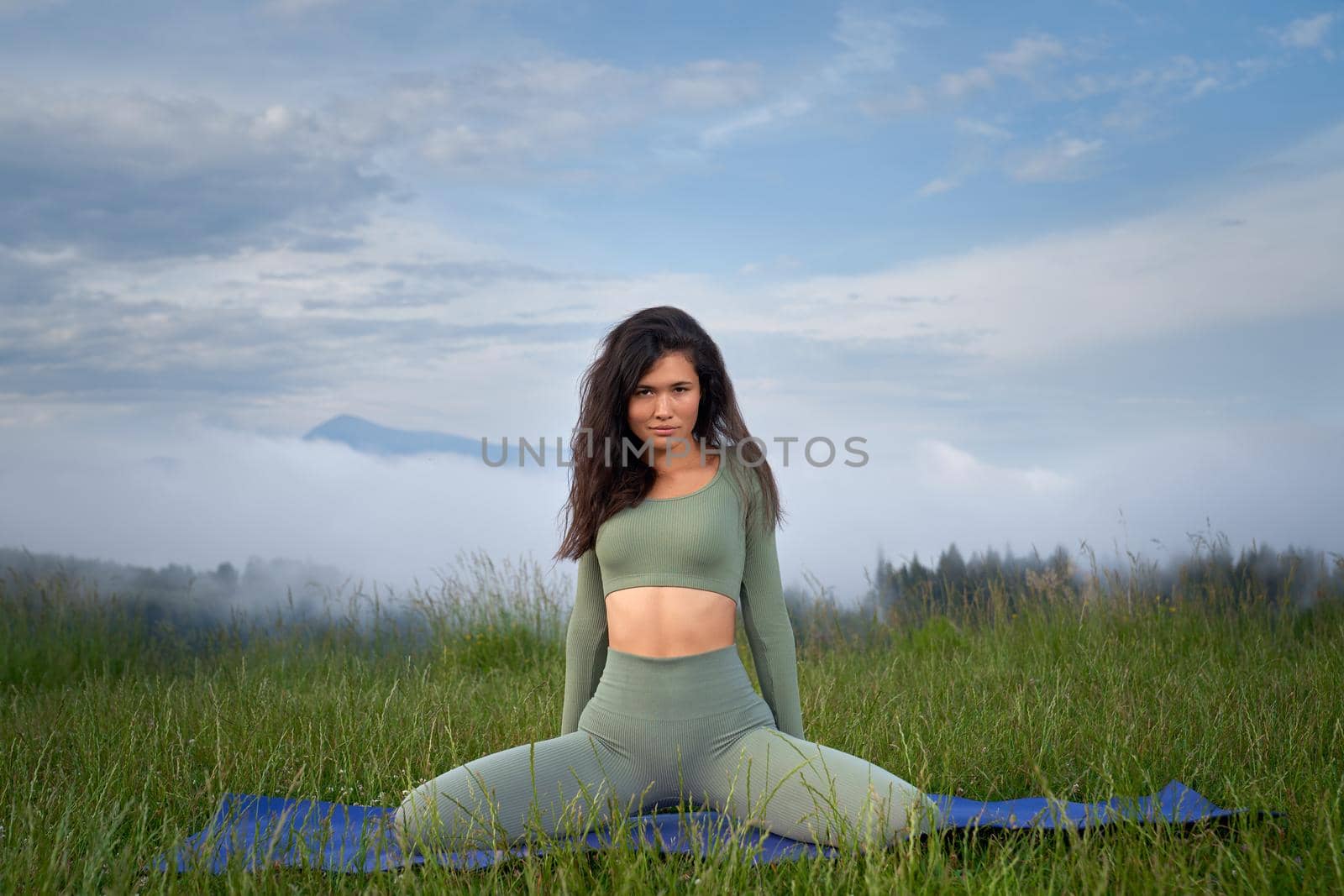 Beautiful young woman with dark hair stretching on yoga mat with mountains on background. Active female person enjoying outdoors activity during free time.