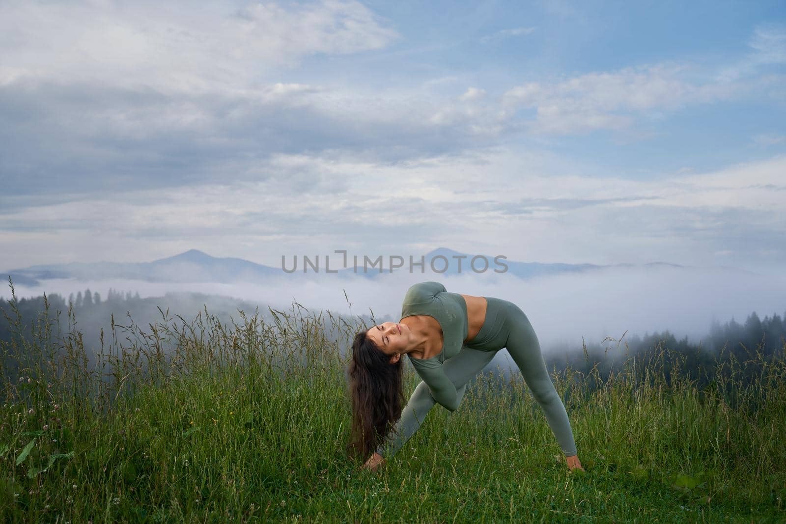 Young active woman with dark hair doing flexible exercises for body among summer mountains. Healthy and fit lady in sport clothes enjoying morning workout on fresh air.